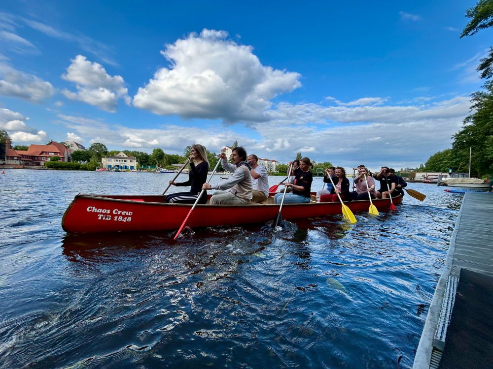 Ein Team paddelt gemeinsam in einem roten Drachenboot mit dem Schriftzug „Chaos Crew“ auf einem Fluss, bei sonnigem Wetter und unter blauem Himmel.