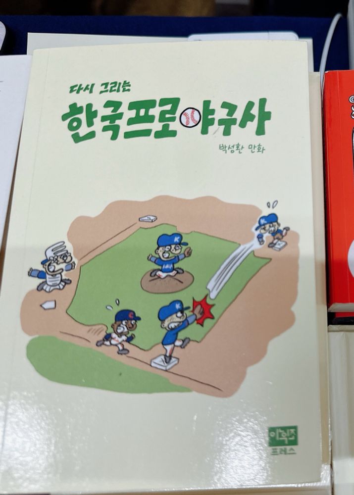 <한국프로야구사> 책. 표지는 명랑만화풍 그림으로, 야구장 내야에서 1루수가 포수에게 공을 던졌고, 주자가 홈에 들어오는 중이다.(아웃된 상황)