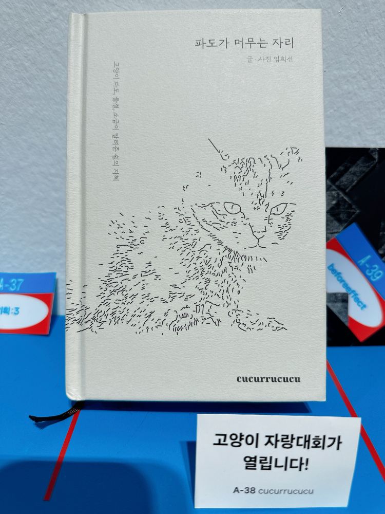 <파도가 머무는 자리> 책. 표지에 펜으로 그린 고양이 한 마리가 노려보고 있다. 책 앞에는 “고양이 자랑대회가 열립니다!”라고 쓰여있는 명함 크기의 종이가 세워져 있다.