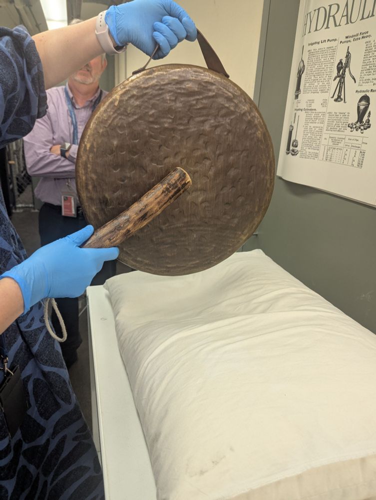 Library staff members striking the brass gong with the wooden striker. Staff member is wearing blue nitrile gloves and holding the gong above a white pillow. Another staff member in the background of the shot is watching and leaning against the wall.
