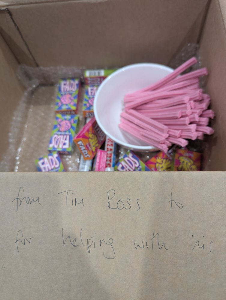 Looking down into a medium sized cardboard box. I side the box are various colourful packets of sweets, including a large amount of pink sticks.