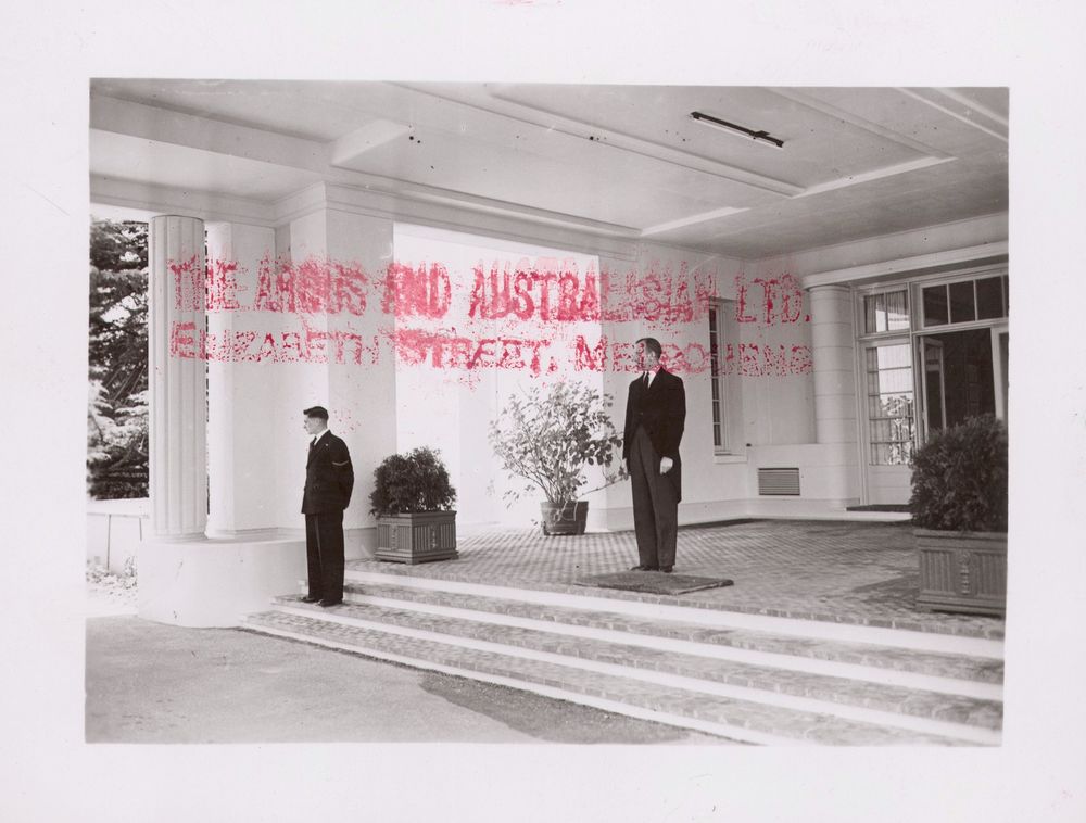 a black and white photo of two men standing on a very large porch. Both men are dressed formally in black attire. The photo print has been stamped with a very large red stamp which reads "The Argus and Australasian Ltd, Elizabeth Street Melbourne."
