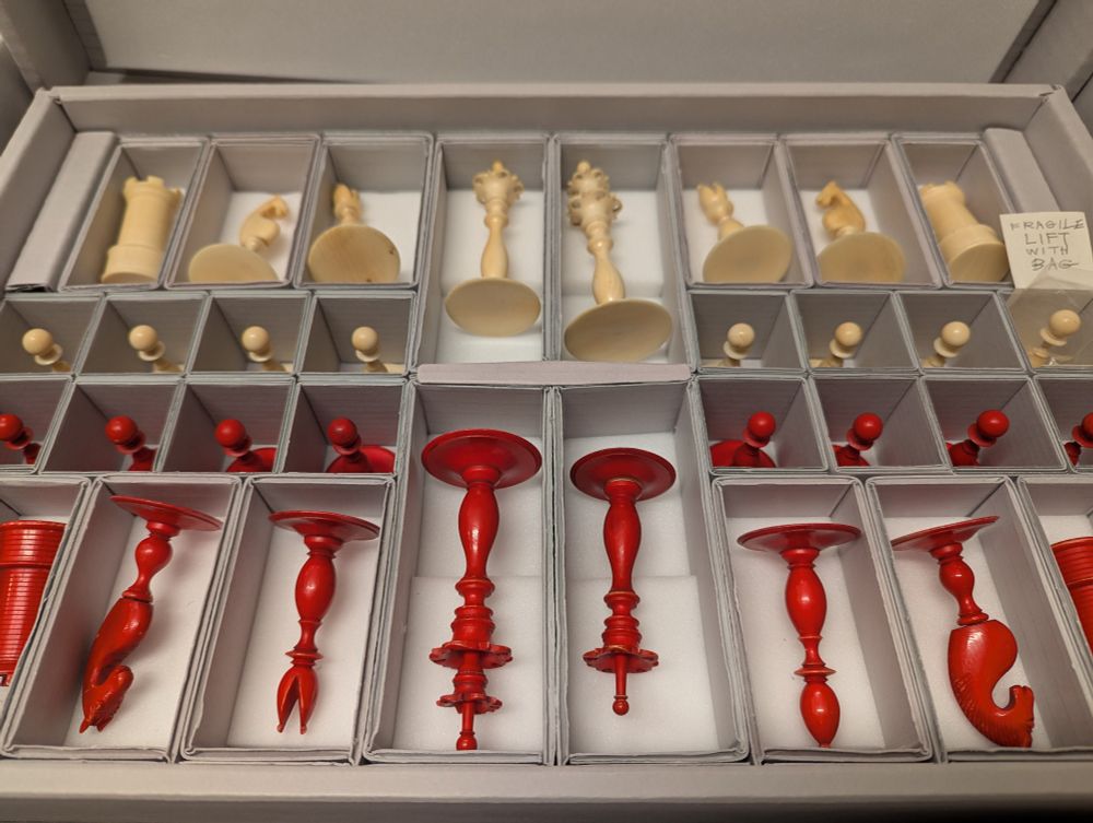 A photograph of chess pieces. The top two rows are a creamy white colour and the bottom two rows are red. The pieces are individually boxed in grey boxes with white padded undersides. They are positioned in four rows. Some pieces are larger than others and are positioned lying flat, whilst some pieces are positioned standing.