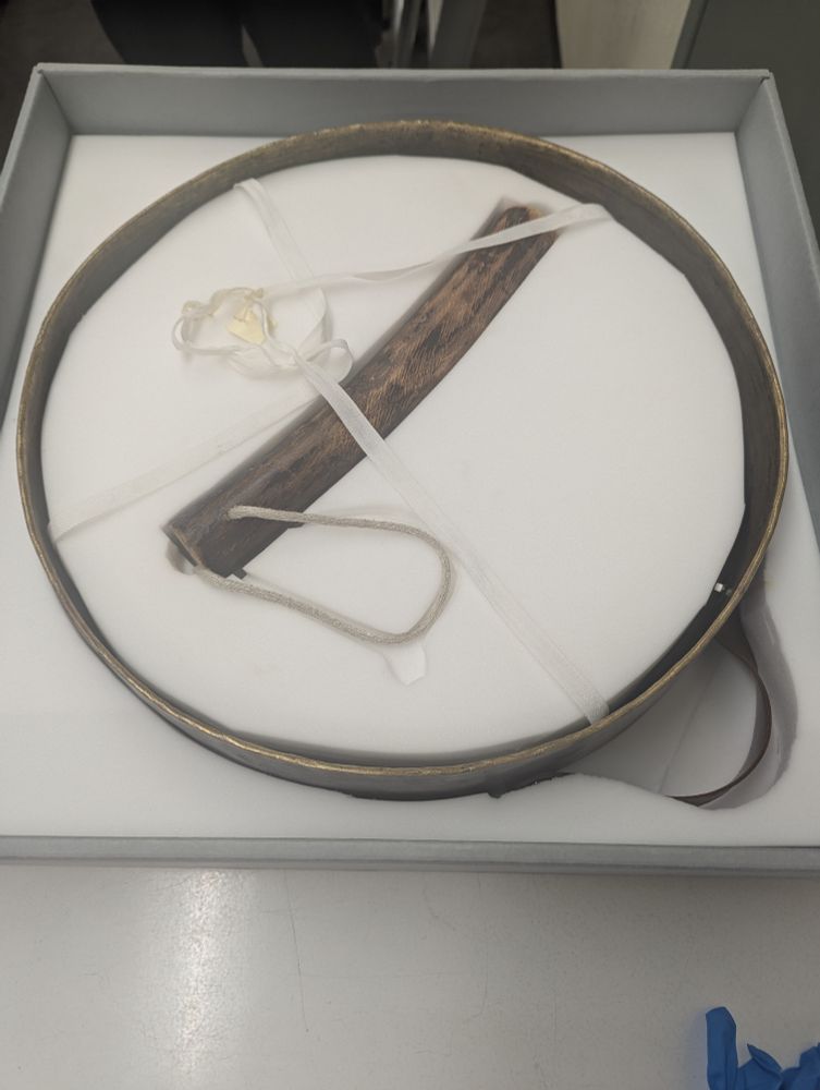 Brass gong laid flat in a box with white padding inside it. A wooden striker is displayed inside it.