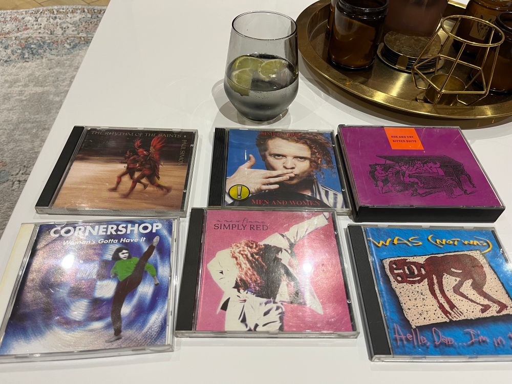 CDs laid out on an Airbnb coffee table with a G&T idling behind them. The CDs are Paul Simon's Rhythm Of The Saints, Cornershop's Woman's Gotta Have It, Simply Red's Men And Women and A New Flame, Hue And Cry's Bitter Suite/Remote and Was (Not Was)'s Hello Dad... I'm In Jail.