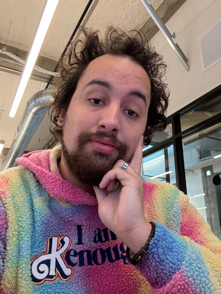 A lightskinned man with a beard poses with his head on his left hand, while wearing a tie dye sweatshirt with the phrase “I am Kenough”.