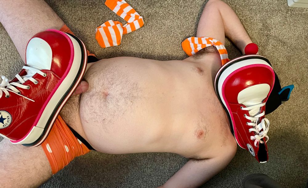 Puppy laying on the floor pretending he’s being stepped on by clown shoes. One is stepping on his hard crotch, the other is on his head. 