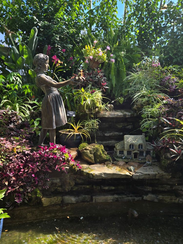 A statue of a girl and a small model of a cottage in a fountain surrounded by orchids