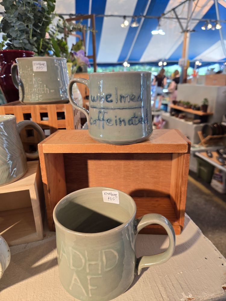 Two light-green mugs, one on a wooden stand. The upper one reads "whelmed caffeinated" with a volume-level bar between the words; the lower reads "ADHD AF"