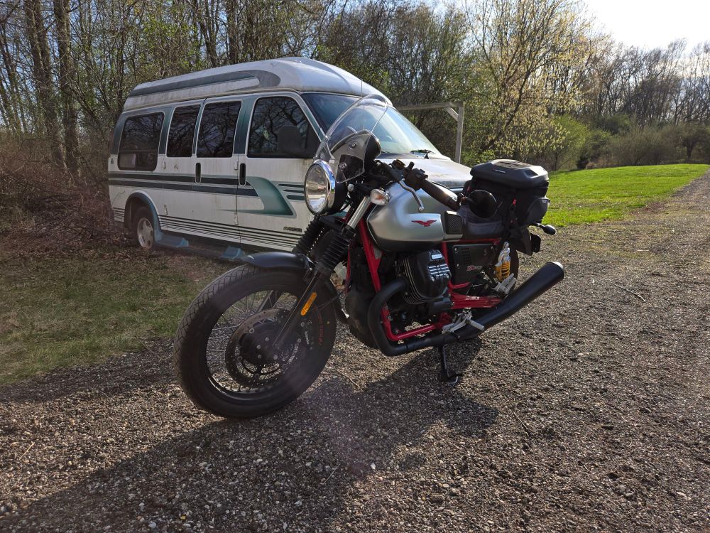 A silver-and-red Moto Guzzi motorcycle in front of a white van, both parked by grass and gravel