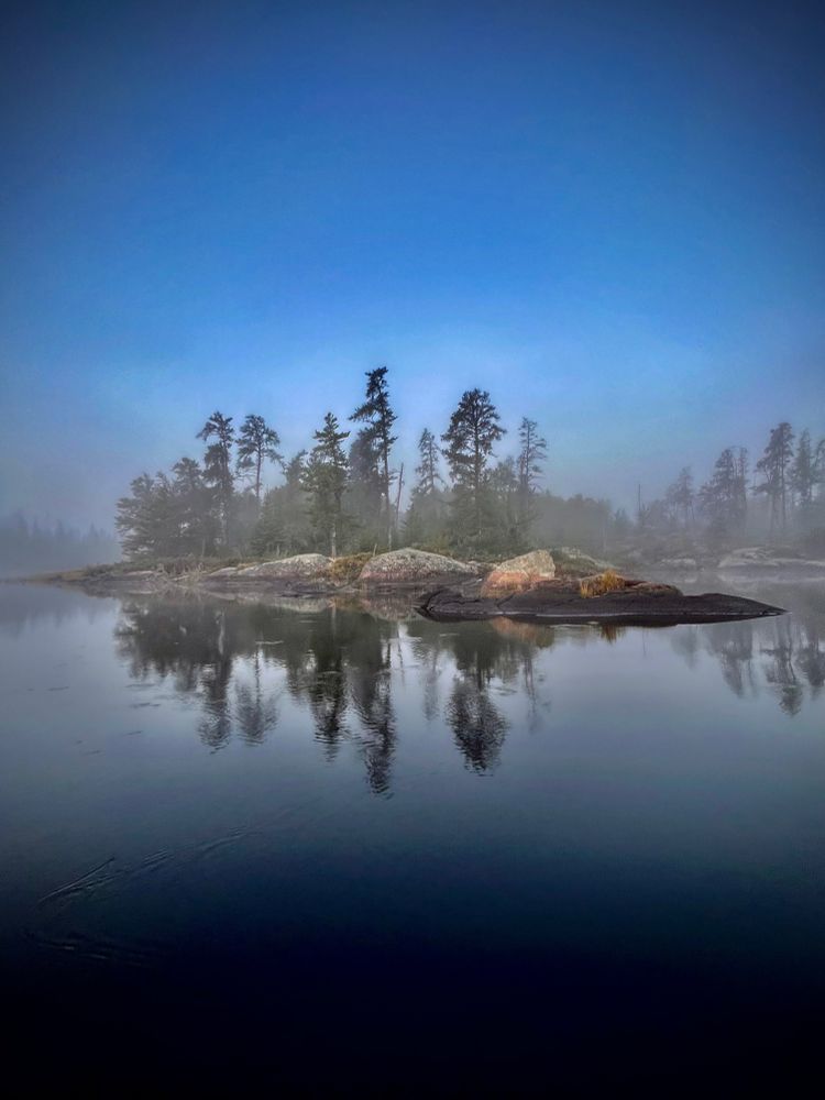 Foggy reflection of a pine trees on a small rocky island on a calm lake. 