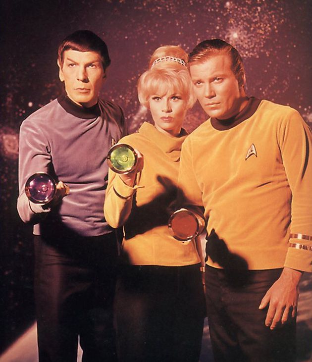Leonard Nimoy in early Spock cut and makeup. Grace Lee Whitney without her signature basket weave hair and wearing a pilot uniform. William Shatner is next to Whitney. Him and Nimoy wear early prototypes of the TOS uniform with rounded color, no jersey cut sleeves and pilot braiding. All hold flashlights with colored backings like futuristic weapons. 
