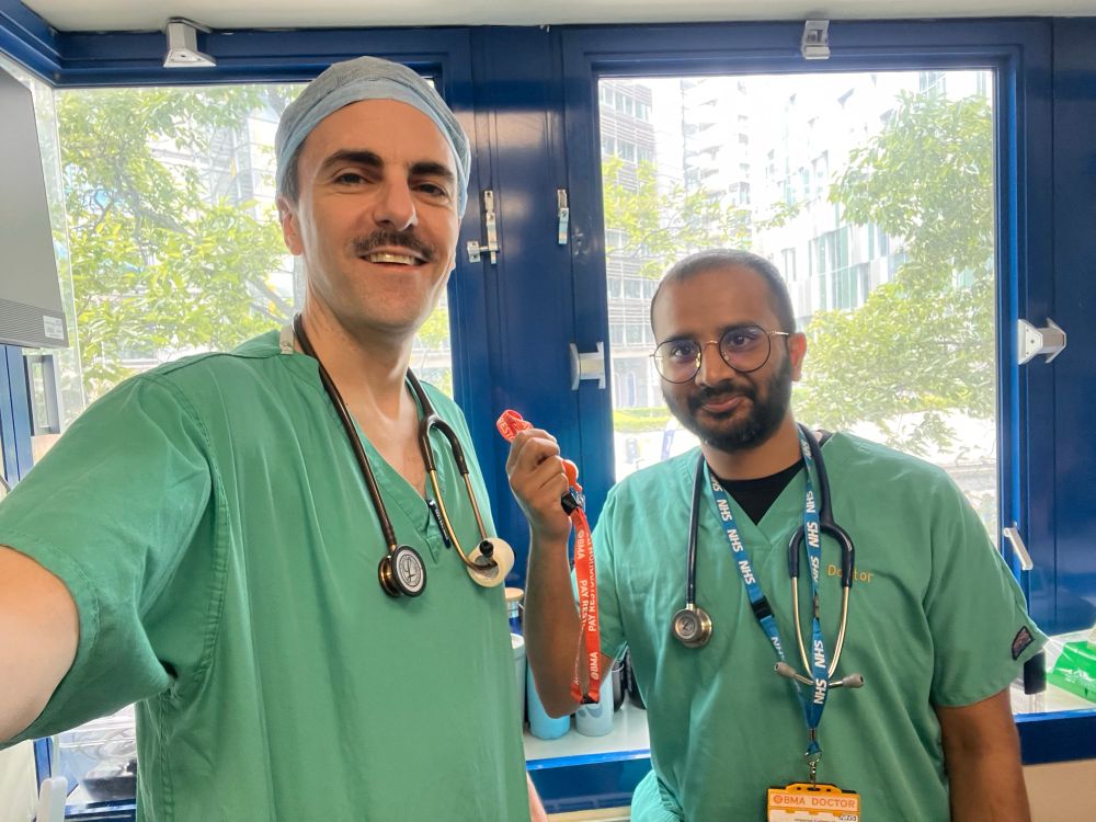 Selfie of Harsha and me, in green scrubs, in the ED office - Harsha is holding up his new BMA lanyard and we’re both smiling like we know we’re going to win this fight