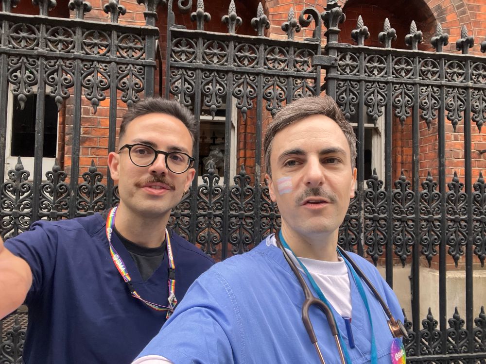Vassili Crispi and Tom Dolphin standing in front of some wrought-iron railings, both wearing scrubs with rainbow accessories