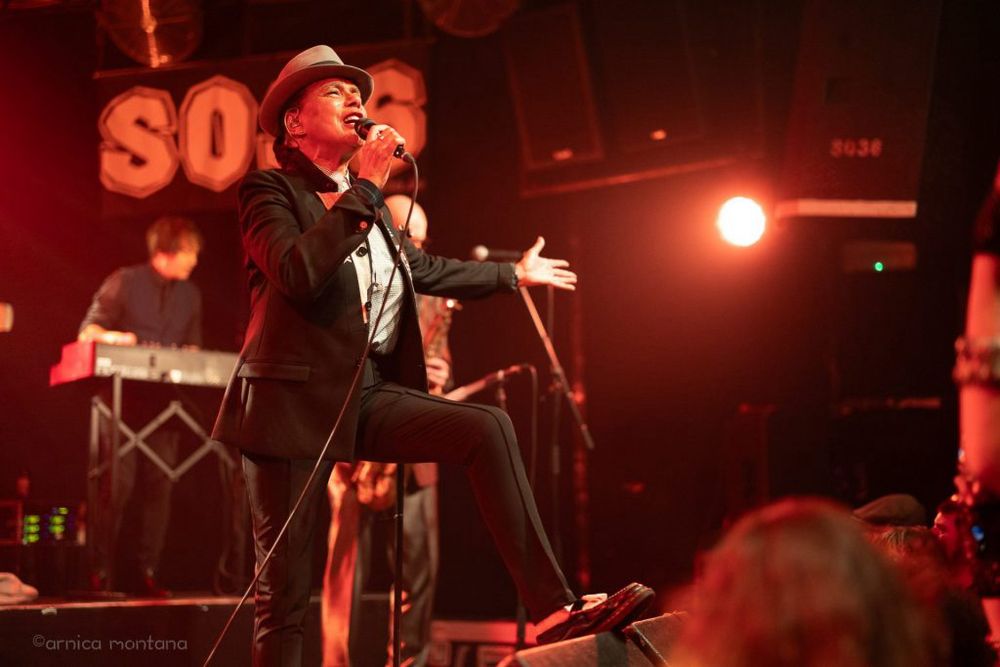 Eine Künstlerin in Anzug und Hut singt auf der Bühne leidenschaftlich in ein Mikrofon, während im Hintergrund ein Keyboarder zu sehen ist. Die Szene wird von rotem Licht beleuchtet, das eine energiegeladene Atmosphäre schafft. Die Zuschauer sind nicht vollständig sichtbar, aber ihre Köpfe sind zu erkennen.