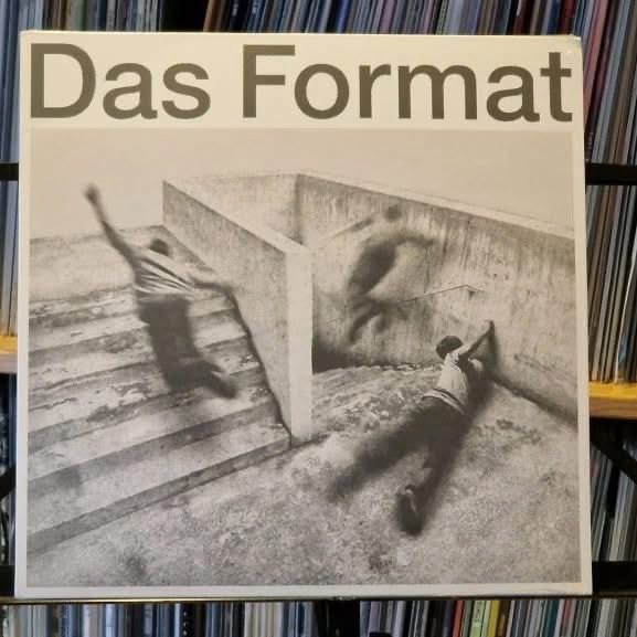 Cover eines Albums mit dem Titel "Das Format". Es zeigt ein Schwarz-Weiß-Bild von zwei Personen in Bewegung, die mit einer konkreten Struktur vor einem Hintergrund von Schallplatten interagieren.