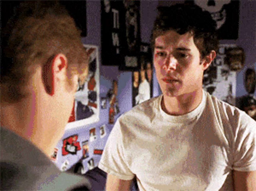 a man in a white t-shirt is talking to another man in a bedroom