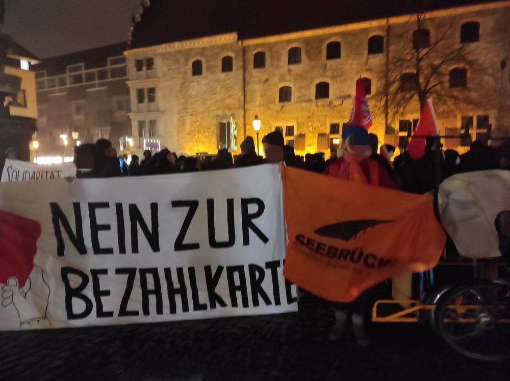 Foto einer größeren abendlichen Menschenansammlung auf einem diesigen, beleuchteten Platz. Auf einem Transpi steht groß: Nein zur Bezahlkarte. Zu erkennen ist auch eine Flagge der Seebrücke.