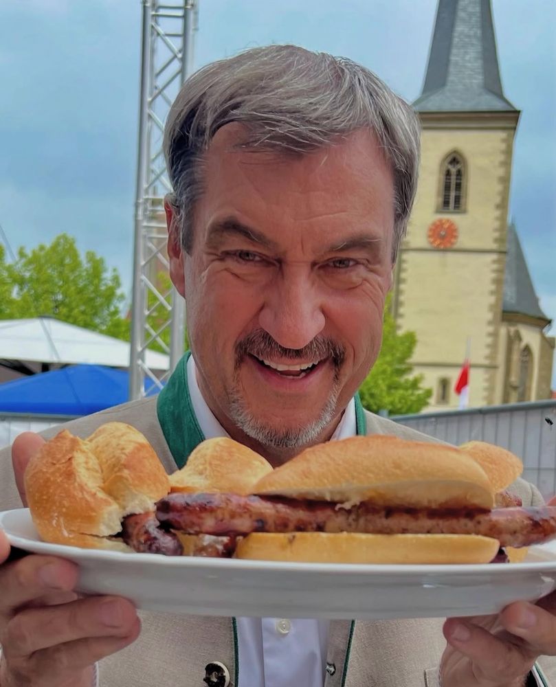 Markus Söder steht im Mittelpunkt des Fotos. Er trägt einen dunklen Anzug mit weißem Hemd und einen leicht geöffneten Kragen, kein Schlips. In der linken Hand hält er eine weiße Papierserviette, in der eine große, gebräunte Bratwurst liegt. Söder beißt gerade herzhaft in die Bratwurst und schaut dabei leicht nach unten. Im Hintergrund sind verschwommen mehrere Menschen zu sehen, teils in Tracht. Die Szene spielt offenbar auf einem Volksfest oder einer ähnlichen Veranstaltung. Das Bild vermittelt eine betont bodenständige, volkstümliche Atmosphäre.