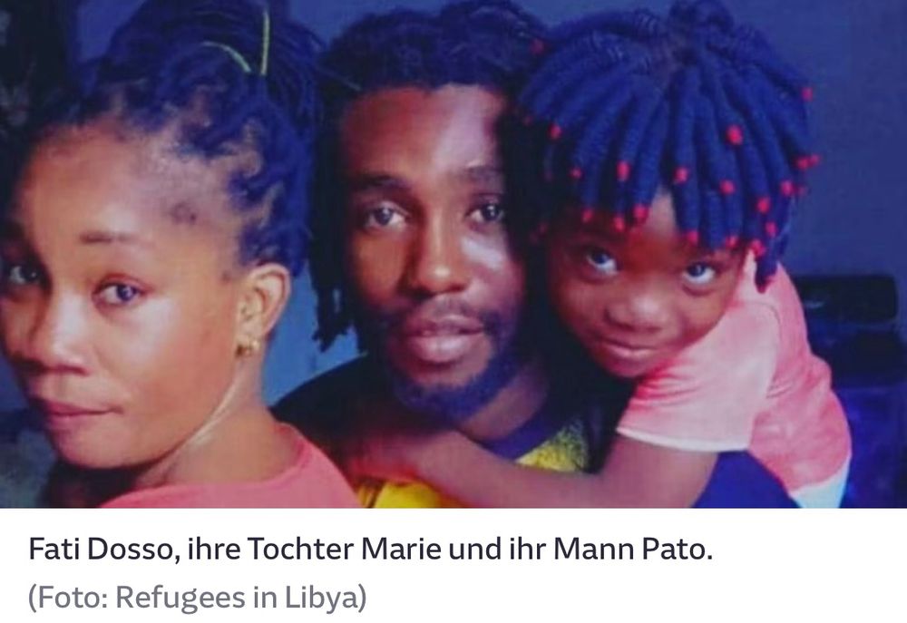 Das farbige Foto zeigt Fati Dosso, ihre Tochter Marie und ihren Partner Pato nebeneinander. Fati steht links im Bild und blickt ruhig in die Kamera. Pato ist in der Mitte zu sehen, mit Bart und offenen, schulterlangen Dreadlocks. Die kleine Marie sitzt rechts auf Patos Rücken und hält sich mit beiden Armen an ihm fest. Sie schaut mit großen Augen direkt in die Kamera und lächelt leicht. Ihre Haare sind kunstvoll geflochten, dunkelblau mit roten Spitzen. Alle drei tragen farbenfrohe Kleidung. Das Foto strahlt Nähe, Verbundenheit und Würde aus. Es stammt von der Initiative „Refugees in Libya“.