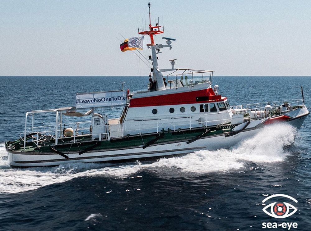 Das Seenotrettungsschiff Sea-Eye 5 auf dem Meer. Das weiß-rote Rettungsschiff der Organisation Sea-Eye fährt mit hoher Geschwindigkeit über das offene Meer. Auf dem Schiff ist ein großes Banner mit der Aufschrift „#LeaveNoOneToDie“ angebracht. Hinten wehen zwei Flaggen: die deutsche Flagge und die Flagge der zivilen Seenotrettung. Das Wasser spritzt am Bug des Schiffs, das dynamisch durch die Wellen pflügt. Unten rechts im Bild befindet sich das Sea-Eye-Logo – ein stilisiertes Auge mit Rettungsring in der Pupille und der Schriftzug „Sea-Eye“.