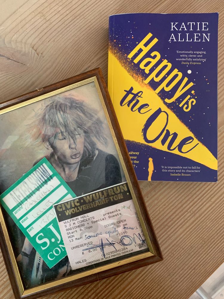 A framed picture of Thom Yorke and gig ticket signed by him to "someone equally famous", the frame is next to the book Happy is the One