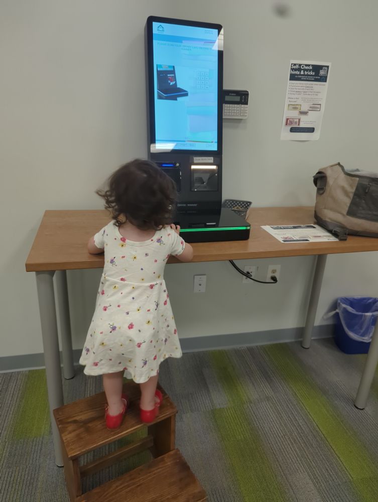 Toddler standing on a step stool in front of a library check out computer. 