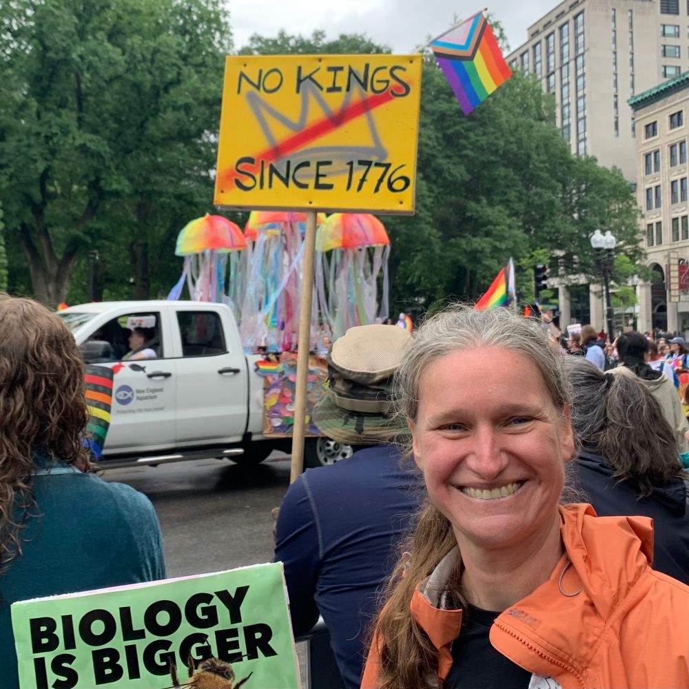 A woman holding a sign that reads Biology Is Bigger stands in a crowd in front of a truck in a parade with rainbow jellyfish and another sign reads No Kings Since 1776.