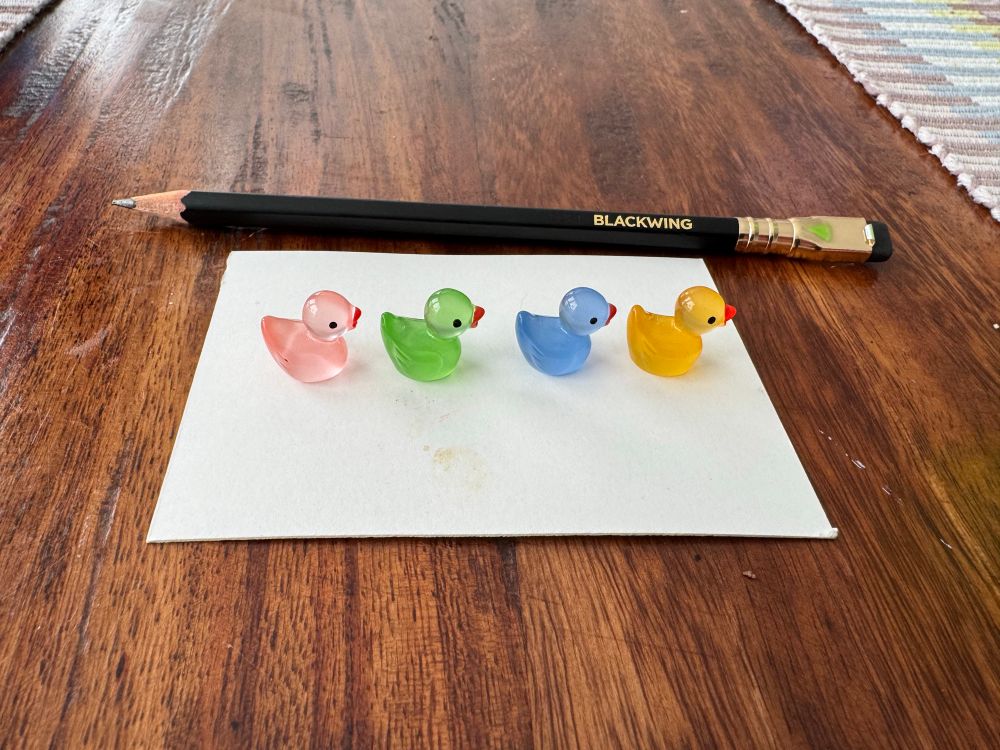 4 tiny acrylic ducks and a drawing pencil gives scale to them. Each duck is about the size of a blueberry