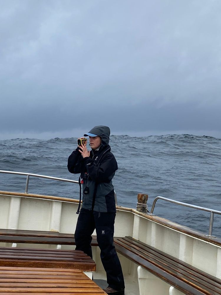 A colour image of a person standing on a boat holding a camera, with the swell of the sea behind copyright Orea Anderson