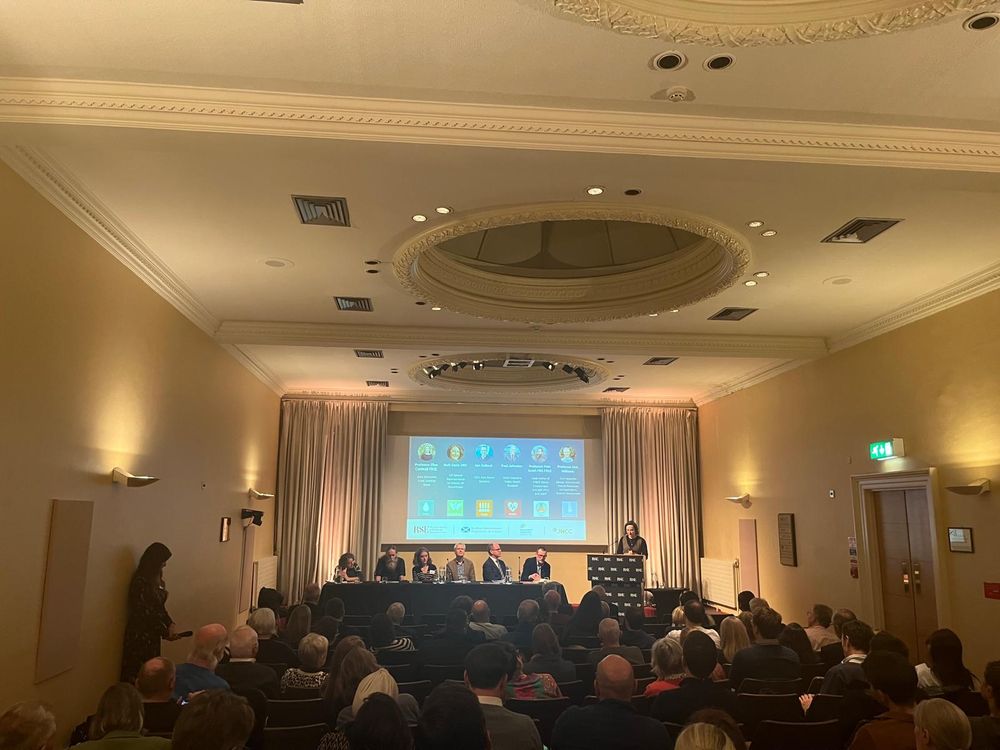 Image shows a full room of people sitting in front of a stage with a speaker stood on the right