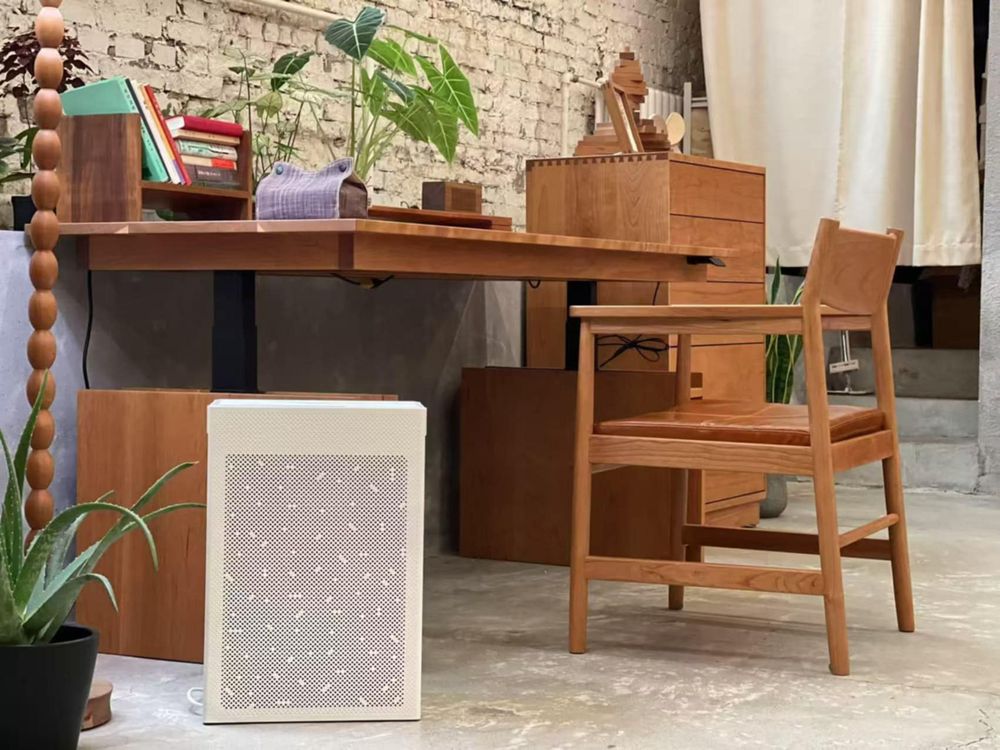 A modern workspace featuring a wooden desk and chair with plants placed on and around the desk. A white SA600 air purifier is positioned on the floor beside the desk, blending with the natural wood and brick wall background for a clean, minimalist look.
