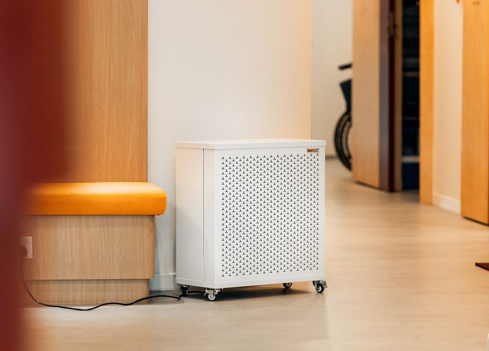 A white air purifier on wheels placed in a hospital or clinic hallway next to a wooden bench with a yellow cushion.