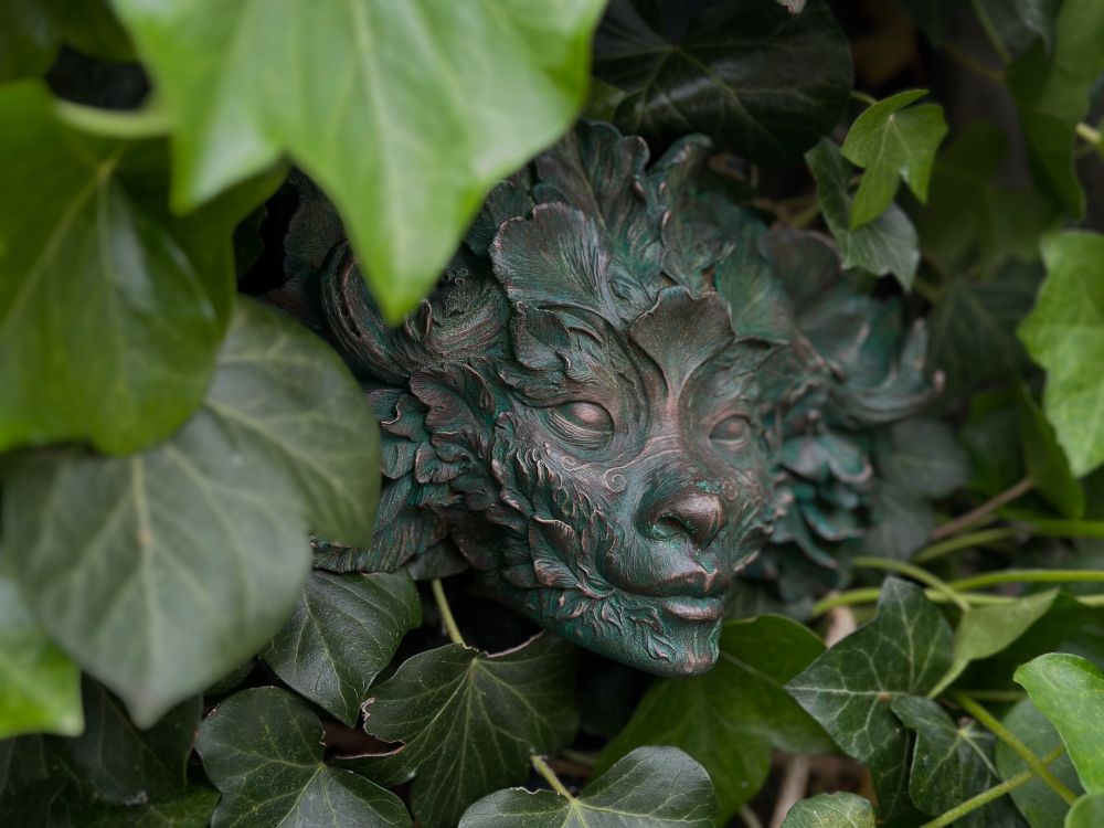 Cold cast bronze sculpture of a green woman on a backdrop of ivy leaves peeking through. Her face is covered in different leaf shapes and swirling scars  
