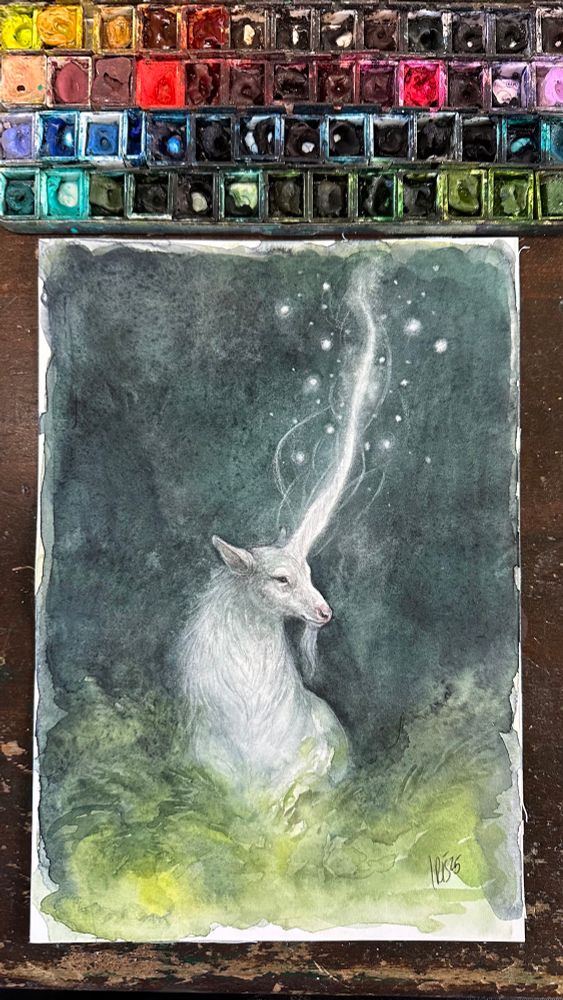 Watercolor painting of a white unicorn stag standing in green foliage against a dark background with motes of light surrounding the glowing horn. 
