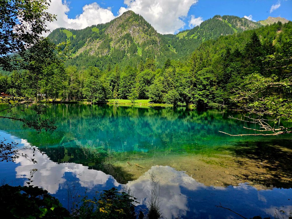 Daylight view of mountains with a beautiful green/turquoise little lake in front. Trees surrounding the lake, which are alo mirrored within the lake