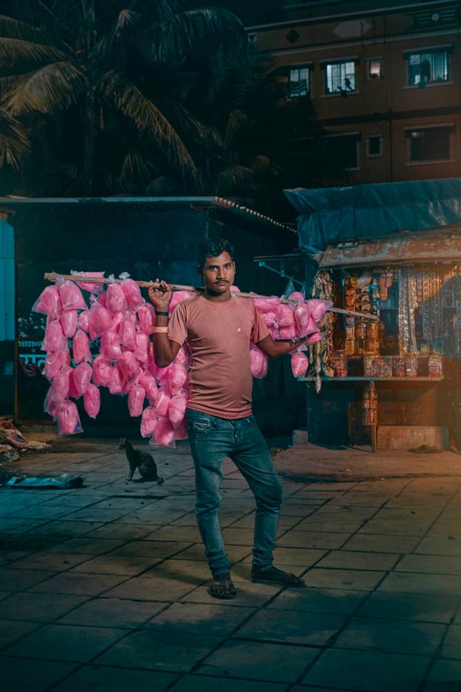 A man has a large stick across both shoulders and it’s laden down with hanging bags of bright pink candy floss 