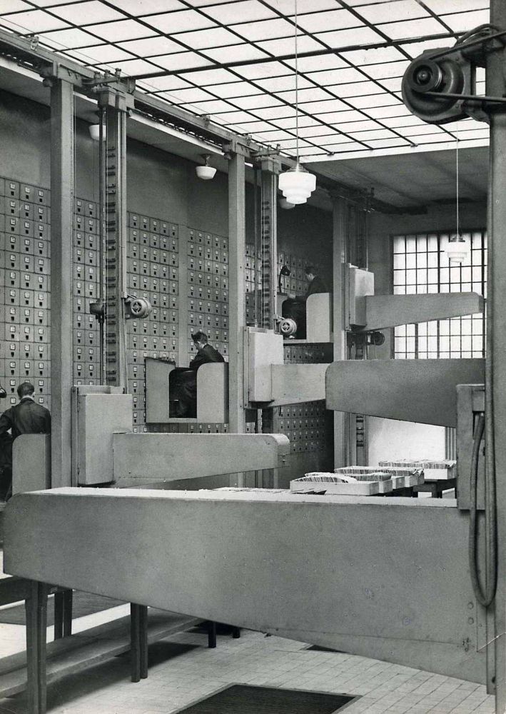 a floor to ceiling wall of filing cabinets, a perfect grid. In front of it are pulley based systems and attached to that three desk cubicles. The ceiling and windows are also grids. 