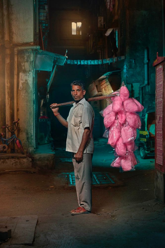 A man stands and over his shoulder is a stock, from it hang bright pink bags of candy floss 