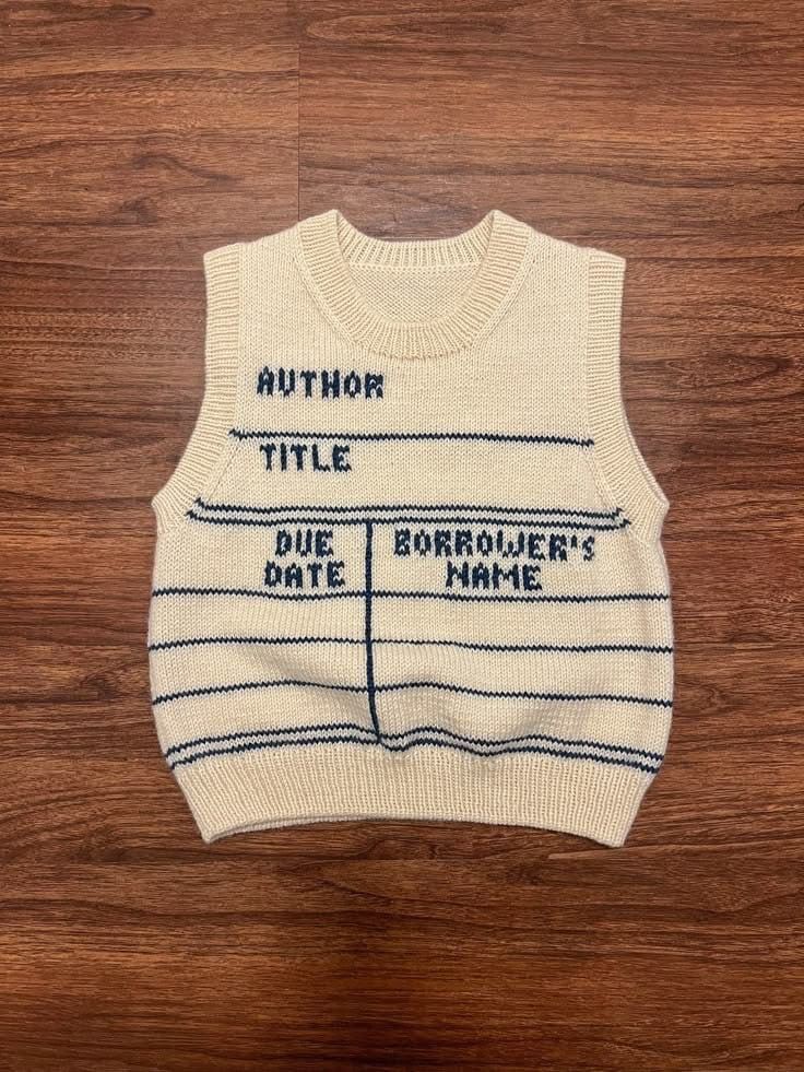 A cream knitted tank top which has blue rules and then author, title, due date and borrowers name also knitted . It’s a library card 
