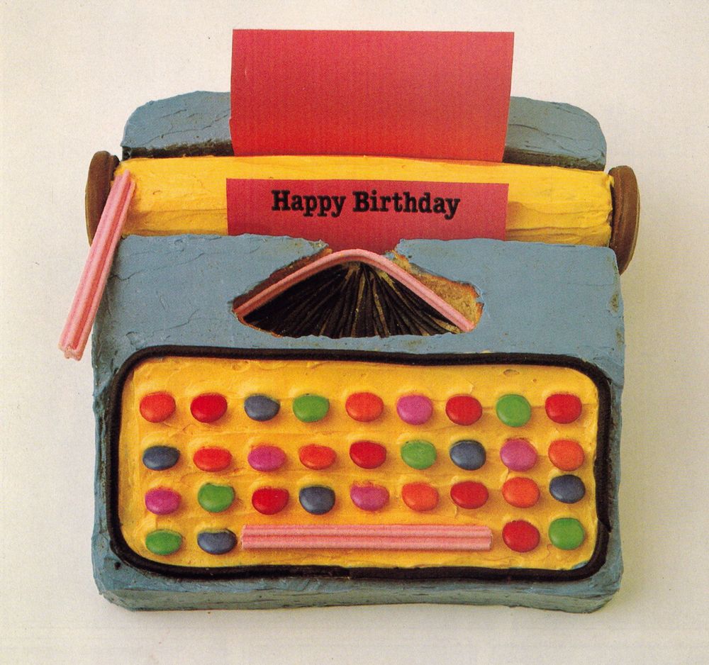A blue typewriter cake, with Smartie keys and a yellow keyboard panel and paper roll. A red sheet of paper sits in it and says Happy Birthday 