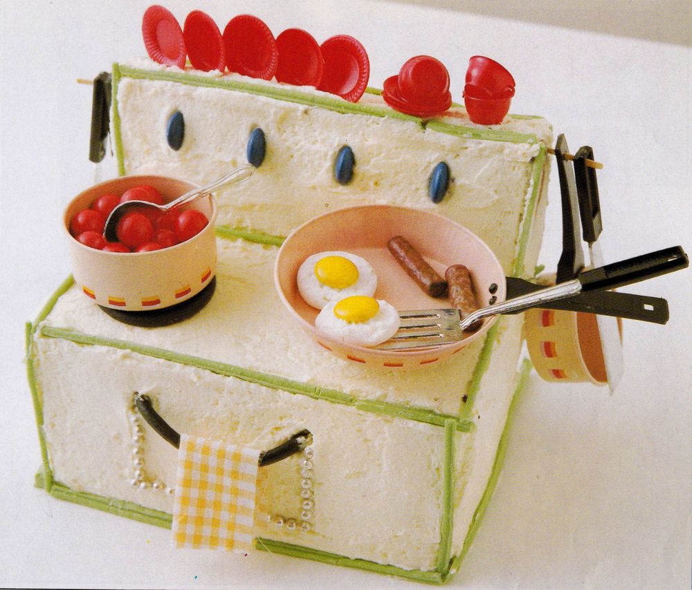 A detailed cake of a stove top and oven. It has two pans on it housing eggs, sausages and red fruit. The cake is iced white with green piping detail. Smarties are placed vertically and pushed into the cake to form controls.