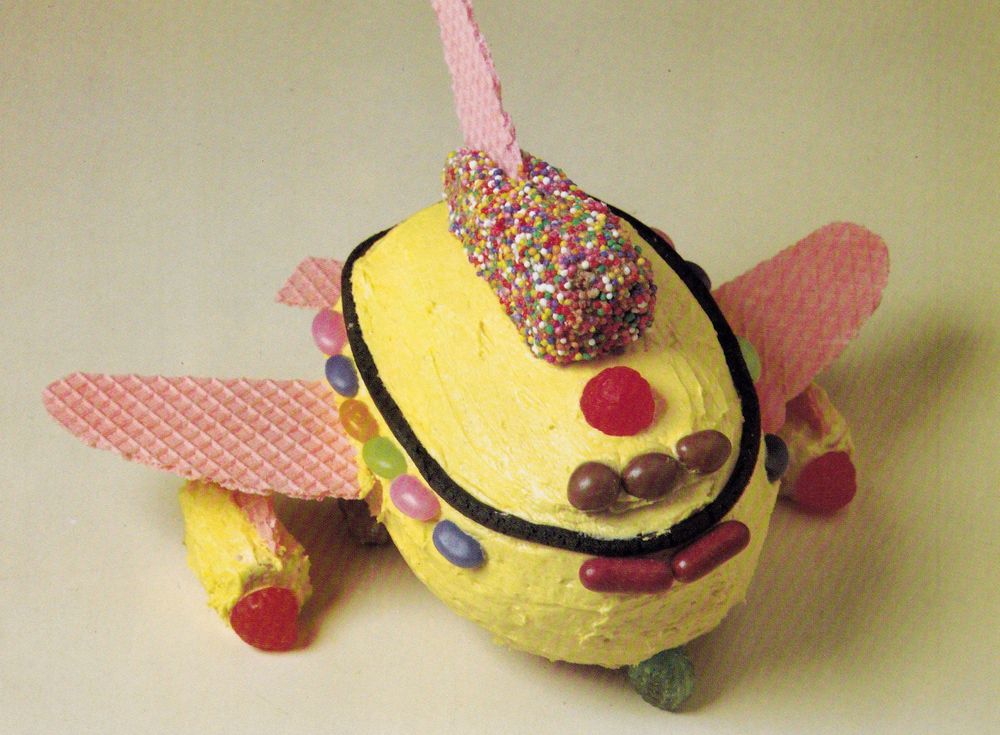 A cake that looks like a chunky, squat aeroplane. It has jellybean windows, pink wafer wings and a yellow buttercream coating. 