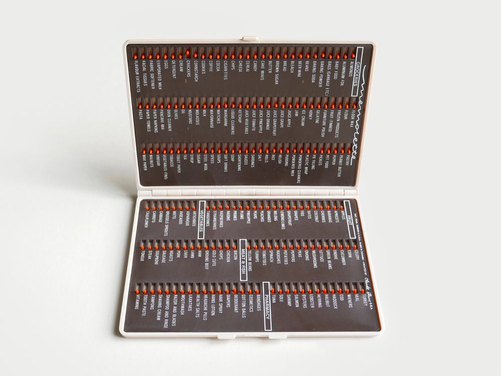 A plastic book like structure showing as open, inside is a dark brown interface that has white type. A thorough list of groceries is written and beside each one is a small orange switch that slides to indicate a purchase.