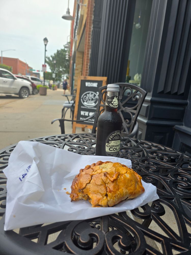 Foreground café table with pasty and soda on it, background downtown small town wirh brick buildings, mud ground Cafe sign advertising tbr Cafe called What the Dickens
