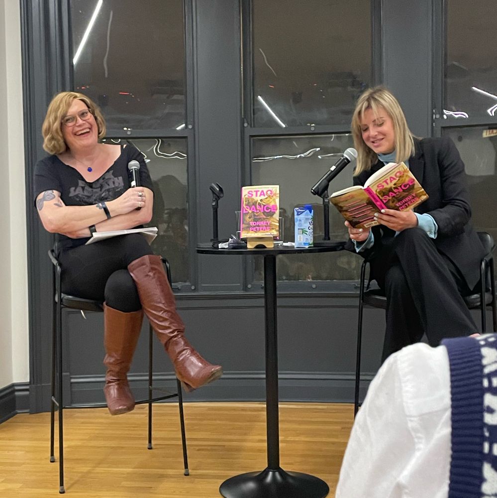 Two black-clad women sit before microphones. One (Torrey Peters) reads from a book entitled STAG DANCE. The other (Lilly Wachowski) chuckles appreciatively. 