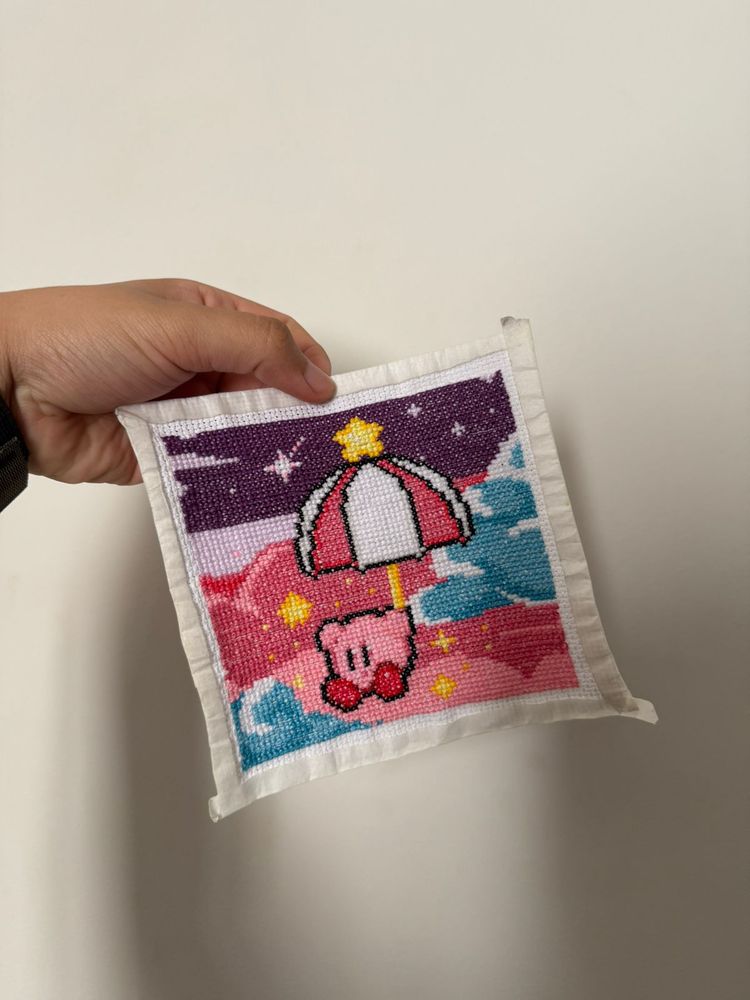 uma foto de um quadrado de tecido costurado em ponto cruz. os pontinhos formam uma imagem do kirby segurando um guarda chuva e descendo pelo céu. o céu é uma mistura de rosa, com nuvens bem azuis e o topo do céu é um roxo escuro com uma estrela. 