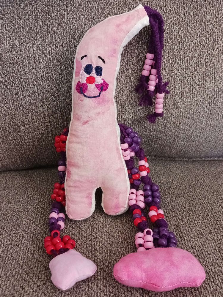 A little pink doll on a couch. They're almost banana shaped but the bottom goes straight down with two stubby legs. They've braided yarn with beads at the ends hanging off the curved tip of their head. They've an embroidered face with two large eyes, a smile, and blushing cheeks. There are two beaded arms with fabric hands on each side of their body.