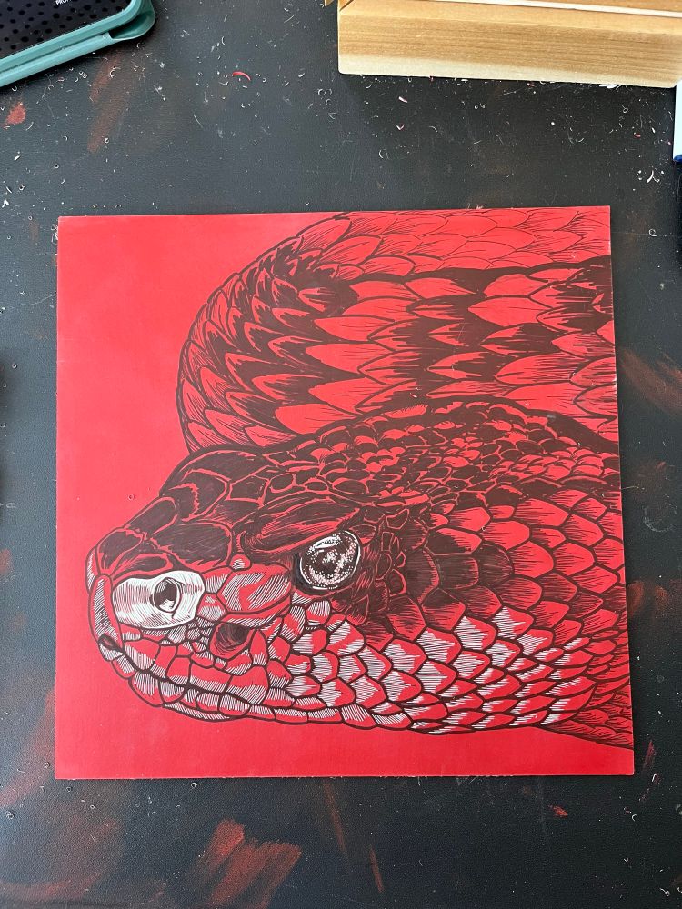 Red linoleum block with a rattlesnake drawn on it. The eye and part of the nose have been carved.