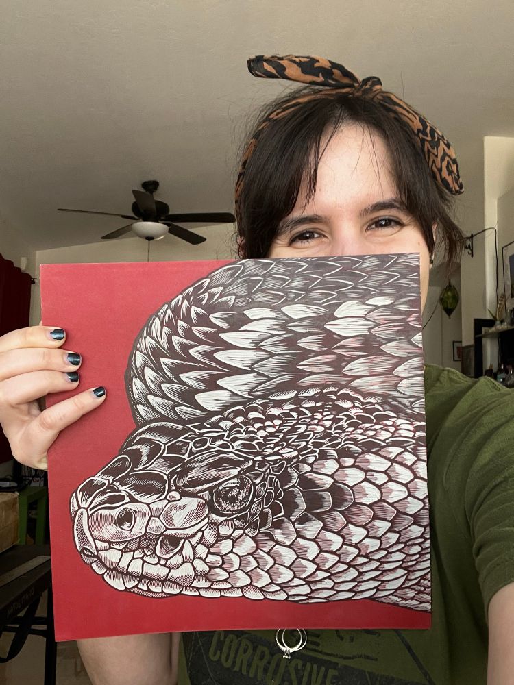 Woman with brown hair wearing a brown bandanna and olive green shirt holds up a linoleum block with a carved drawing of a rattlesnake on a red background on it. The linoleum block partially obscures her face but you can see her eyes smiling. 